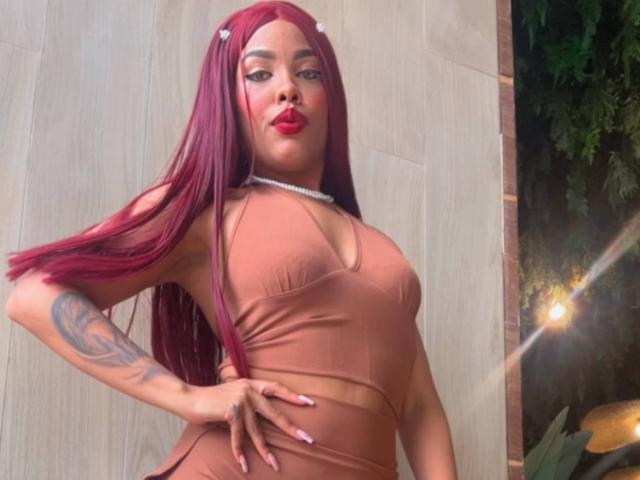 LexieBloom with red hair, wearing a brown crop top and matching skirt, showing a tattoo on her left arm, hand on hip, wearing red lipstick.