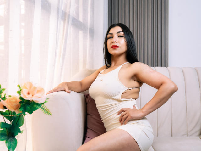 ChanelBrouw with black hair, red lipstick, and a white cut-out dress, sitting on a beige couch, hand on hip, showing her ass and a small tattoo on her arm.