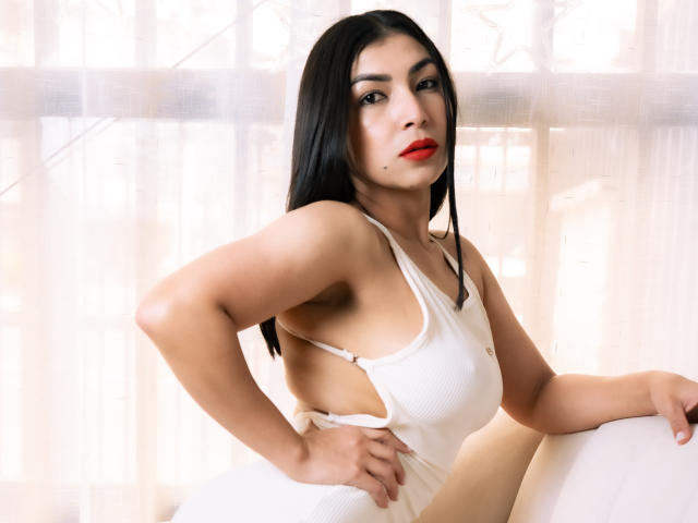 ChanelBrouw with black hair, red lipstick, and white ribbed tank top showing sideboob, hand on hip, sitting on white couch, soft light, confident expression.