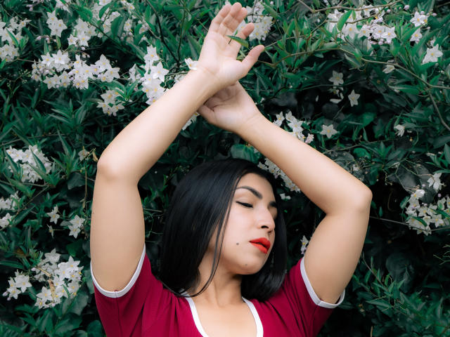 ChanelBrouw with arms raised, dark straight hair, red lipstick, small mole under her nose, wearing a red and white top, standing in front of white flowers and green leaves.
