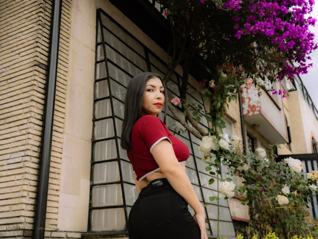 ChanelBrouw has big tits, a cute face, and a sexy ass. She's wearing a red top, black pants, and standing outside with purple bougainvillea and white roses.