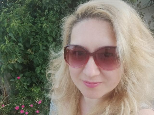 AmaliaDesire with blonde wavy hair, wearing pink sunglasses, pink lipstick, smiling softly, showing her nose and upper chest, outdoors with green foliage and pink flowers.