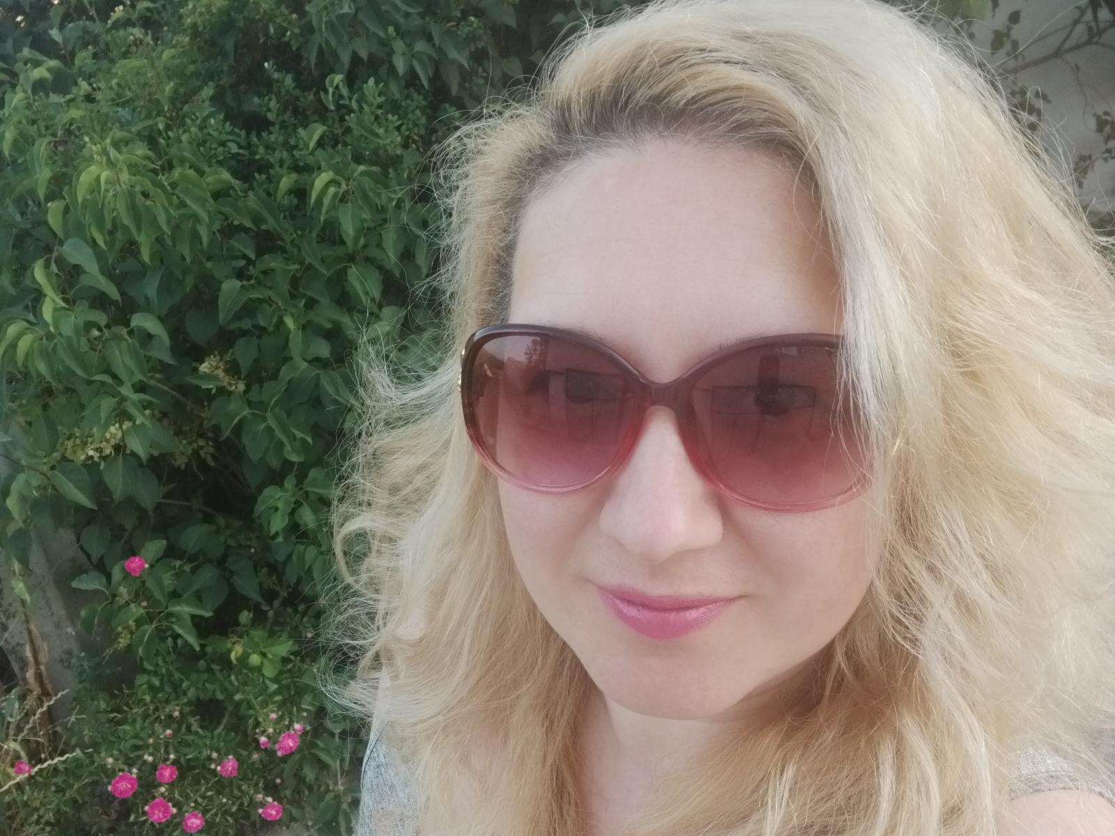 AmaliaDesire with blonde wavy hair, wearing pink sunglasses, pink lipstick, smiling softly, showing her nose and upper chest, outdoors with green foliage and pink flowers.