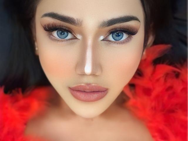 LustHaven's face with blue eyes, full lips, and dark eyebrows, wearing a red feather boa, has a white stripe down her nose. She's looking straight at the camera.