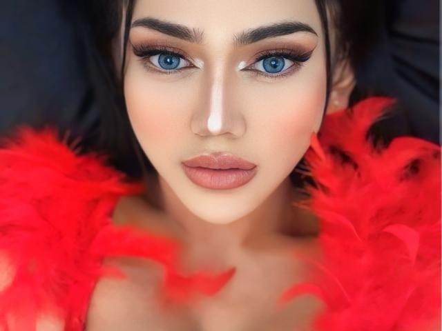 LustHaven has blue eyes, full pink lips, and is wearing a red feather boa. She has dark eyebrows, long eyelashes, and pale skin. Her nose is slightly upturned.