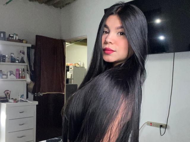 BiancaJeins with long black hair, red lipstick, and dark eyes, looking over her shoulder, showing her ass, in a room with a white dresser and flat-screen TV.