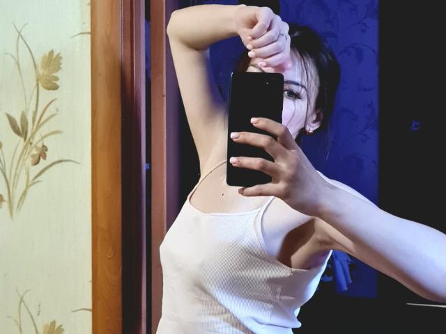 TinaPeachX with pale skin, dark hair in a messy bun, wearing a white tank top, stretching her arm above her head, holding a black phone, mirror selfie.