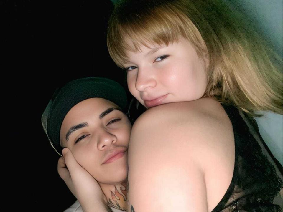 CouplExtreme69 with blonde hair and bare shoulders, leans over a man with tattoos, black cap, and slight smile, against a dark background. Man's nose touches CouplExtreme69's shoulder.