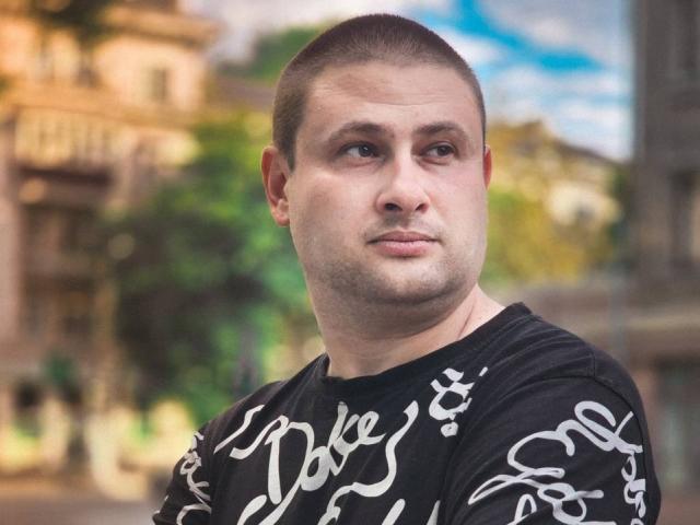 TimDesire, a chubby dude with a buzzcut, wearing a black shirt with white script, looks off to the side with a slight smirk. His nose is small and his face is stubble.