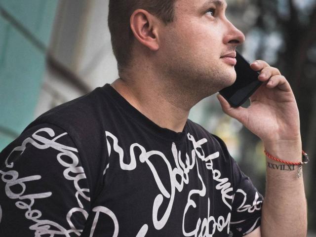 TimDesire with short brown hair, black t-shirt with white script, holding phone to ear, looking to the side, wearing red bracelet with Roman numeral III.