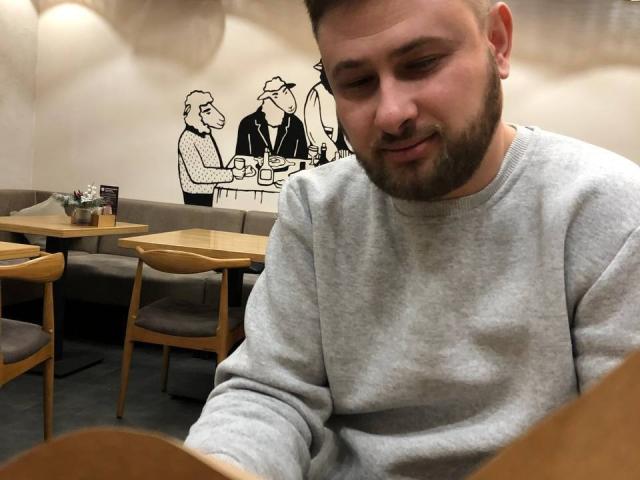 TimDesire, a bearded dude with a light brown beard and short hair, wearing a gray sweater, sitting in a cafe with wooden chairs and a wall art.