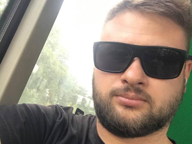 TimDesire has a beard, wearing black sunglasses, and a black shirt, with short brown hair, looking slightly annoyed. His green earbuds are visible.