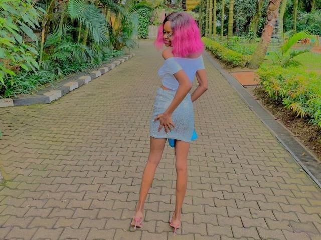 AlexaZane with pink hair, small tits, tiny blue skirt, white top, hands on ass, standing on brick path, wearing high heels, greenery in background.