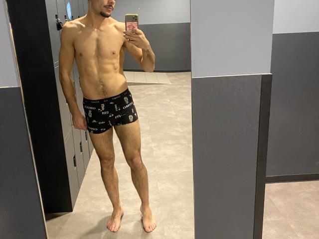 KaelXl69, senza camicia, indossa slip neri con motivi bianchi, in piedi di fronte a uno specchio, tiene in mano un telefono, muscoloso, pelle chiara, capelli corti e scuri, con un leggero sorriso..