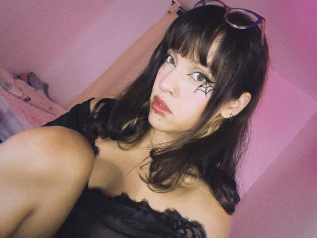 LyaPink with dark brown hair, black off-shoulder top, nose ring, spider web tattoo under right eye, red lipstick, eyeglasses on head, knee up, pinkish light, slightly pouty expression.