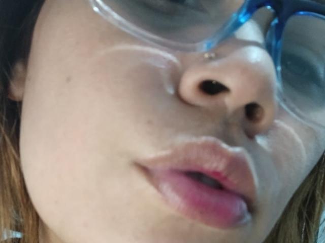 A shot of SophiaQueens' face, showing her nose, pink lips, and blue-framed glasses with a shiny, slick substance on her cheek and nose. Her brown hair is visible.