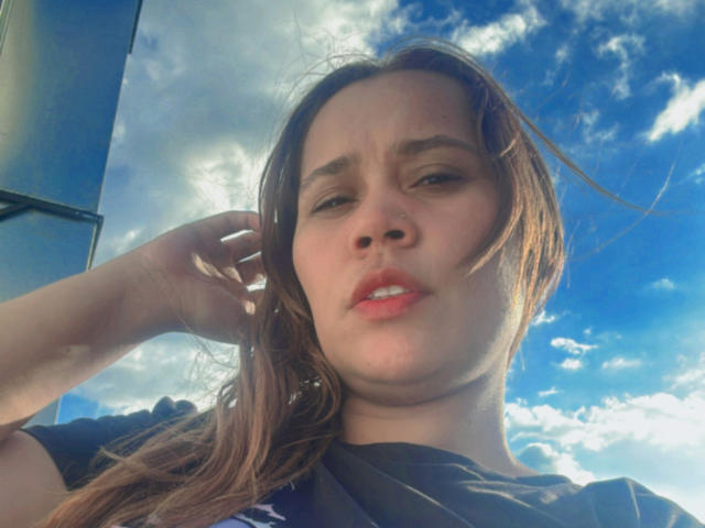 SophiaQueens with puffy red lips, slightly squinted eyes, and brown hair, wearing a black shirt, has a confused or annoyed expression, with her hand in her hair.