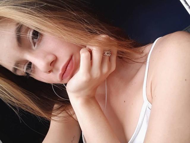 Mahbi with blonde and brown hair, white tank top, hand on cheek, earbuds in, pink lips, ring, slight smile, fair skin, looking at camera, close-up, soft lighting.