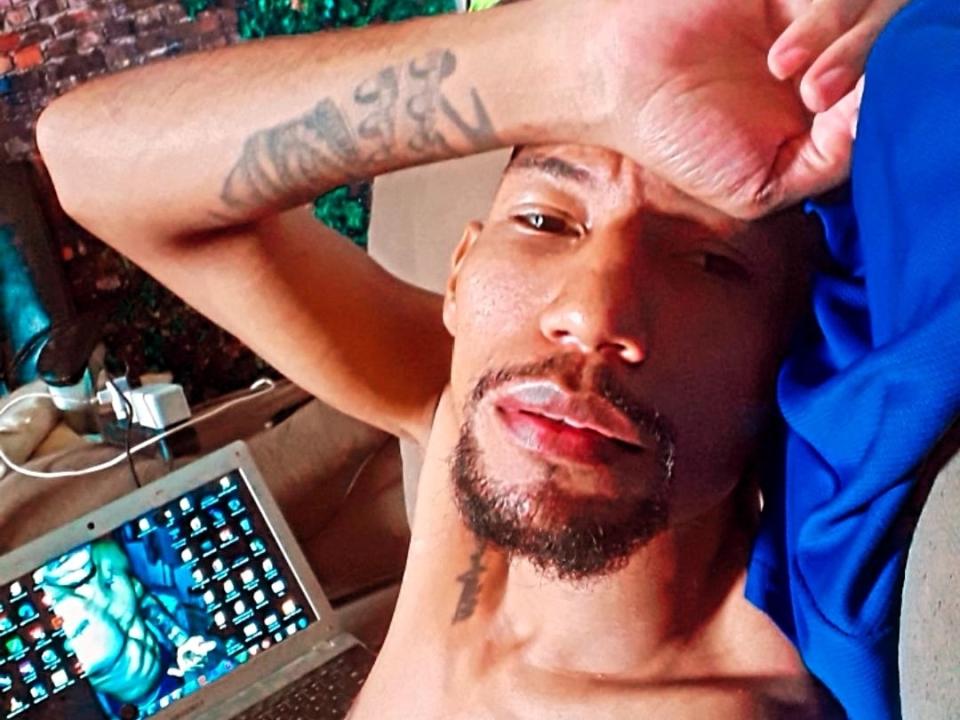 WellJhon with a tattoo on his arm, blue shirt, and a laptop showing a video, lying back with one arm over his head, looking at the camera with a slight smile.