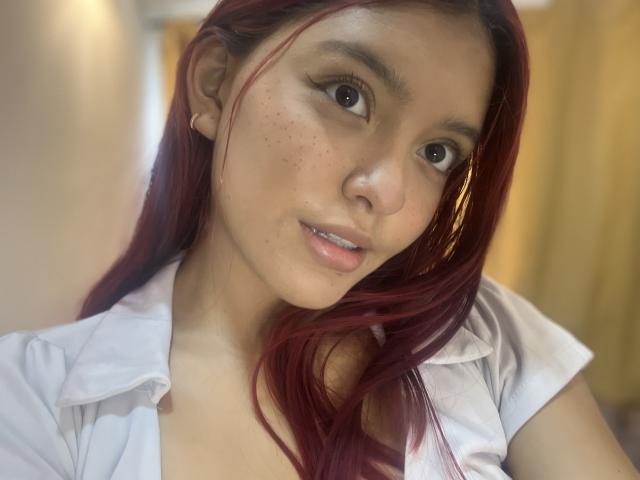 AlyMartiny, avec ses cheveux roux, ses taches de rousseur et son joli visage, porte une chemise blanche et regarde l'appareil photo avec un léger sourire. Elle a un nez mignon et de petites seins..