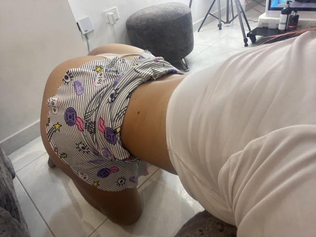 El trasero de AlyMartiny, con ropa interior a rayas y estampados coloridos, inclinada sobre el suelo de baldosas blancas, vistiendo una camisa blanca, mostrando un trasero bronceado, liso y con una pequeña peca..