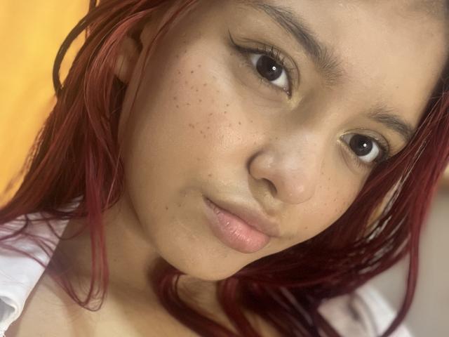 A AlyMartiny tem cabelo ruivo, olhos castanhos e sardas no rosto. Ela está olhando para a câmera com os lábios rosados ligeiramente entreabertos. Sua camisa branca está visível..