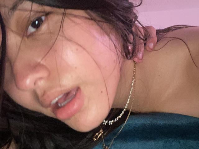 AlyMartiny's face with messy black hair, slightly open mouth, and a gold chain necklace, showing a pinkish cheek and a hand in her hair. Her eyes are half-open.