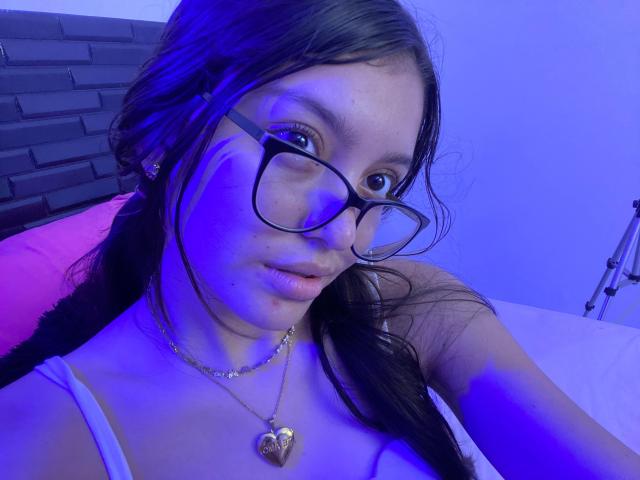 AlyMartiny, com o rosto iluminado em azul, usando óculos, uma camiseta branca e um colar de coração, tem cabelos longos e escuros, e seios pequenos, olhando para cima com um leve franzir de lábios..