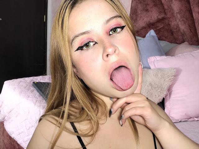 MelaniBecker has blonde hair, green eyes, pink eyeshadow, and is sticking her tongue out with her finger near her mouth. She's wearing a black top with a cute face.