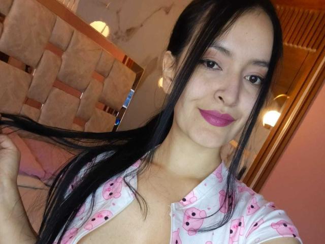 AriaBonnet with long black hair, pink bear print shirt, and pink lipstick, smiling at the camera, holding her hair. Her tits are visible under the open shirt.