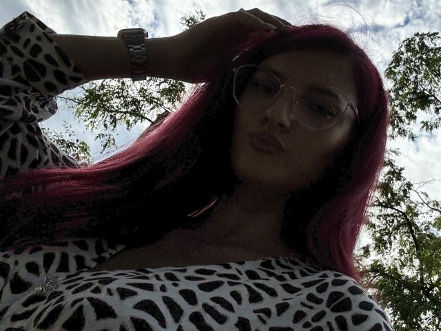 Rosalyn, con cabello rojo, gafas y una blusa de estampado de leopardo, haciendo pucheros con la mano en el pelo, usando un reloj, mirando hacia abajo. Se ven sus senos, con el cielo y un árbol en el fondo..
