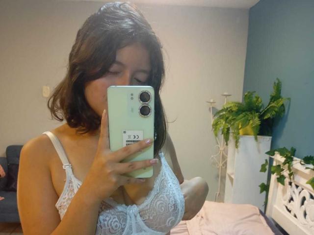 SaraiiAngel with medium brown hair, wearing a white lace bra, holding a green phone, taking a mirror selfie, showing her boobs, in a softly lit room.
