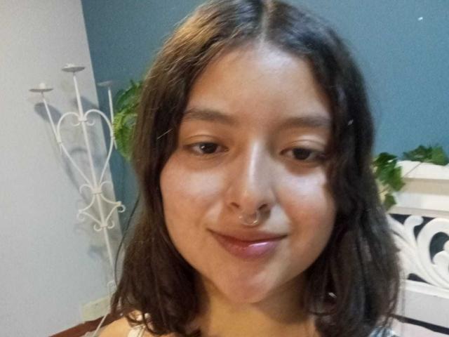 SaraiiAngel has medium brown skin, wavy dark brown hair, a nose ring, and a slight smile. She's wearing a pink top, and her brown eyes are relaxed.