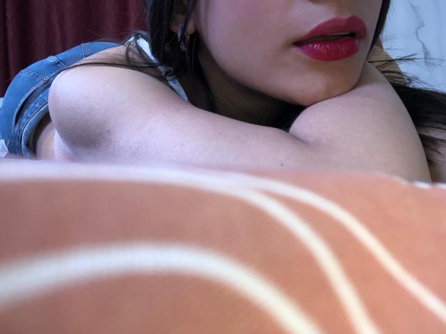 TinyLaurent with red lipstick, arms crossed, lying down, showing her bare shoulders, denim shorts, and a close-up of her orange-striped pillow in the foreground.