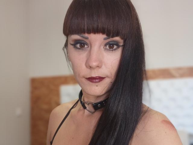 OrianaLaFrancaise has dark brown straight hair with bangs, wearing a black choker with a ring, and has dark eyeliner, red lipstick, and a small mole under her right eye.