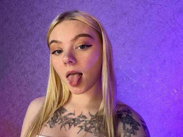 EnchantingEyes with blonde hair, sticking out her tongue, has black eyeliner, tattoos on her chest, and a purple light background. She's topless, showing her small, perky tits.
