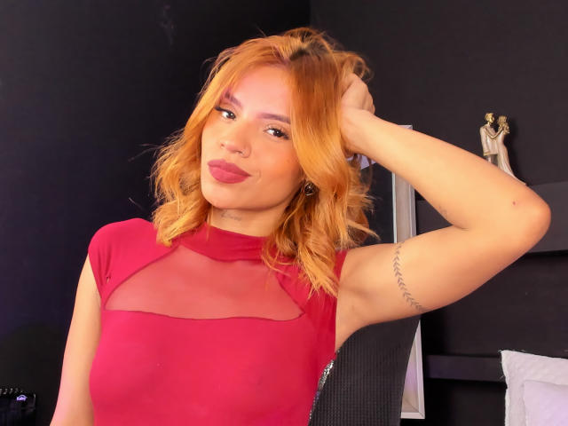 MartinaNovac with orange hair, red sheer top, raised arm, tattoo on right arm, red lipstick, smirking, holding hair, cute, sexy, attractive, feminine, bedroom, soft lighting.