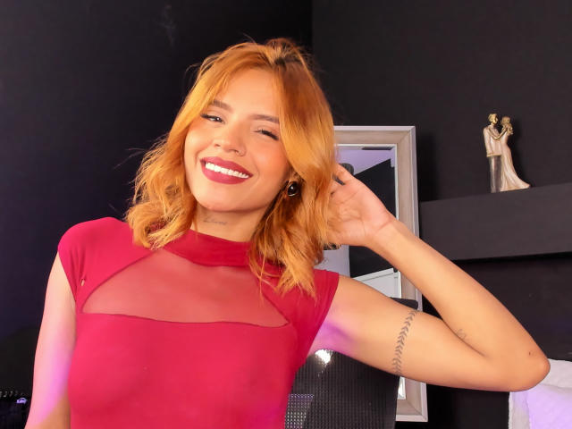 MartinaNovac with orange hair, smiling, wearing a red sheer top, showing her tits, has a tattoo on her right arm, and is winking. She's touching her hair.