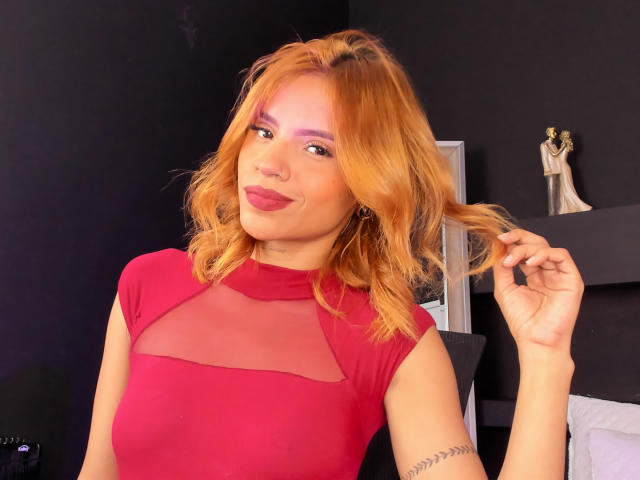 MartinaNovac with orange hair, red sheer top, purple eyeshadow, and red lipstick, holding her hair, showing a tattoo on her arm, smiling at the camera.
