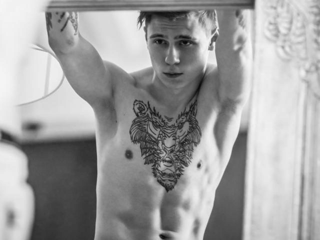 TylerGarner, shirtless, with a detailed tattoo of a lion's face on his chest, arms raised, looking at the camera, black-and-white, muscular, short hair, tattoos on arms.
