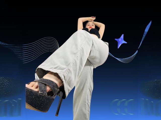 KatherineToro with arms raised, wearing white pants and black sparkly high heels, her large tits and ass prominent, foot in foreground, blue star and wavy lines in background.
