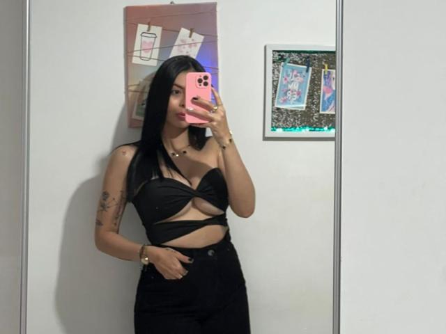 IsabellaJames with long black hair, wearing a black strappy top and black pants, taking a mirror selfie with a pink phone, showing tattoos on her left arm.