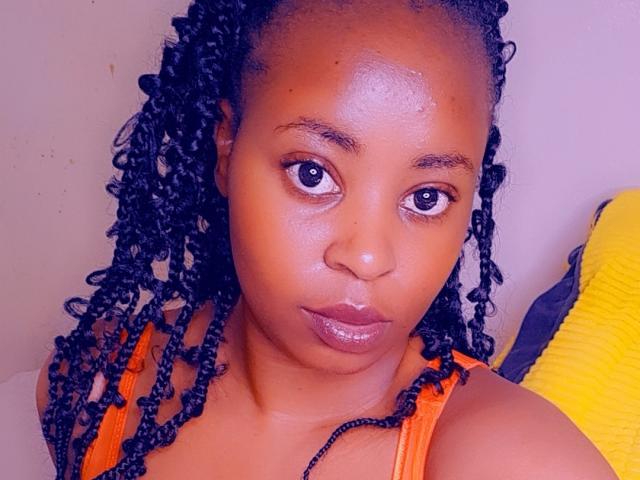 QueenBoobby with dark brown skin, medium-sized tits, and shiny lips, wearing an orange top, has black braided hair, and is staring directly at the camera with a neutral expression.