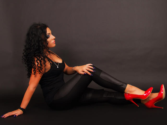 MissKhloe, with curly black hair, sits on the floor in black lace top, shiny black pants, and red high heels, showing her ass and legs, against a dark background.