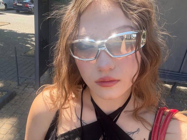 Bubochka with wavy brown hair, wearing shiny reflective sunglasses, black strappy top, and red striped shoulder bag, sunlight reflecting off glasses.