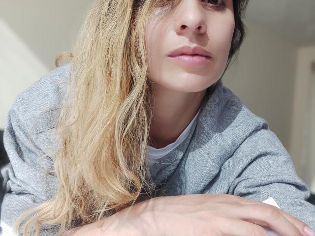 SkylerSamuel has light blonde hair, pink lips, and wears a grey sweater, looking down with a slightly pouty expression, hands crossed in front. Soft natural light.