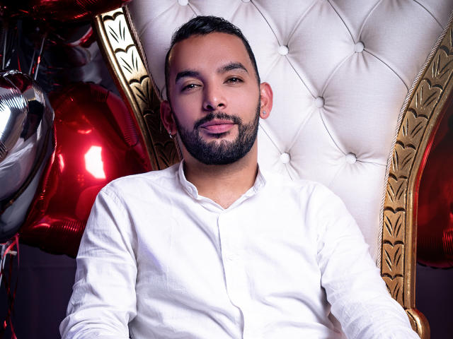 PauloDaSilvaa with a beard, short black hair, and a white button-up shirt, sitting on a tufted, gold-embellished chair, with shiny red and silver balloons in the background.