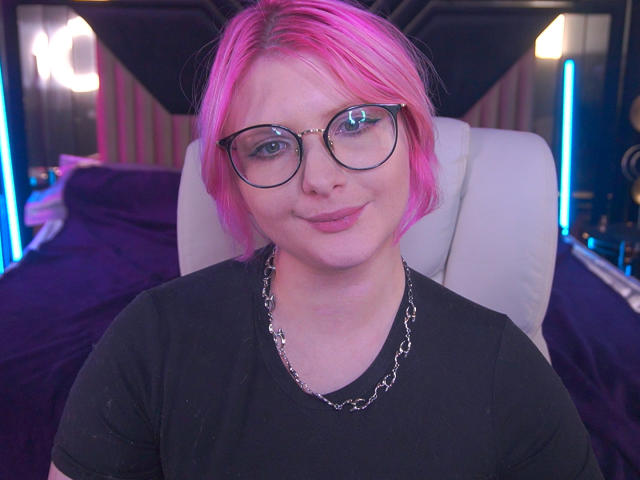 CoolTatis with pink hair, glasses, and a silver chain, wearing a black shirt, smiling slightly, looking cute and nerdy.