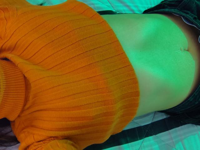 AmyStreetwise69's orange ribbed sweater, exposing her flat stomach and navel, with green lighting on her skin, lying down with dark green pants visible.