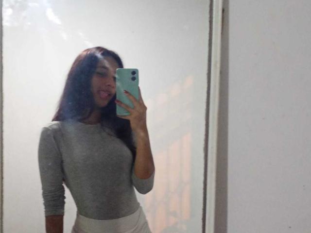 BeautifulIndie with long dark hair, gray long-sleeve top, and white pants, holding a teal phone, taking a mirror selfie, tongue slightly out. Boobs visible under tight top.